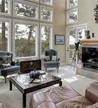 Sunny living room with large windows, two blue chairs, a fireplace, a telescope, a glass coffee table, and a leather couch. Tall trees are visible outside through the windows.